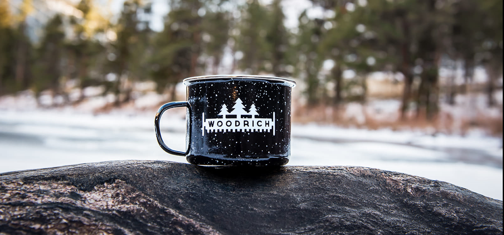 A black coffee mug, garnishing the Woodrich logo in white, placed upon a rock with snowy woods as the backdrop.
