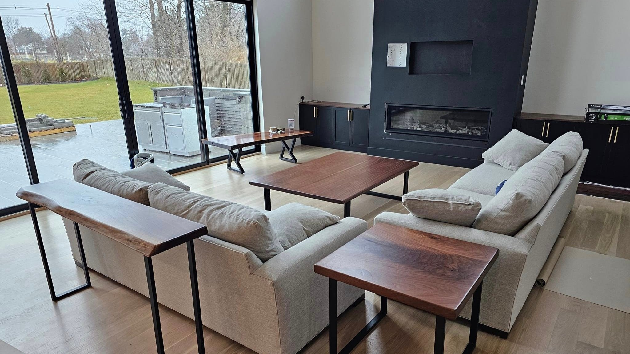 A walnut console table, end table and two coffee tables made with powder-coated black steel legs staged in a modern living room. The furniture shares the room with two light gray couches, a black fireplace and glass sliding doors that lead outside.