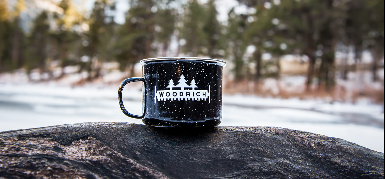 A black coffee mug, garnishing the Woodrich logo in white, placed upon a rock with snowy woods as the backdrop. 